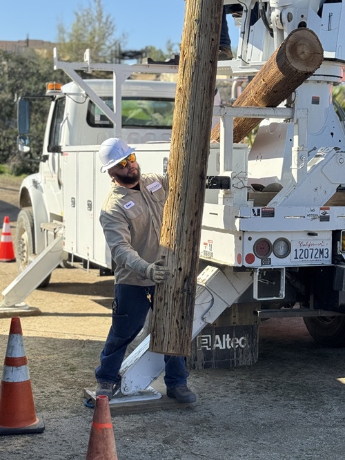 TBB 16 Anza pole unload An Anza Electric Coop utility worker wearing a white construction helmet helps guide a utility pole from off a big utility truck