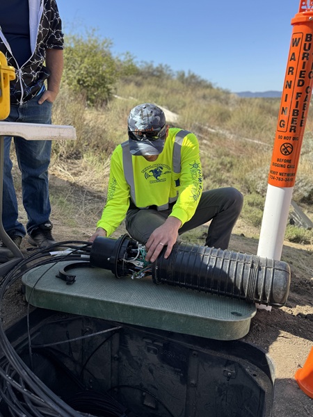 Dustin and splice case vault TBB instructor kneels next to an open fiber vault in the ground examining and exposed cylindrical splice case
