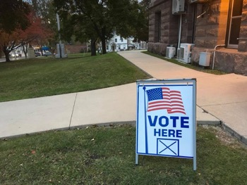A sign that says "vote here" is placed on a lawn outside of city building