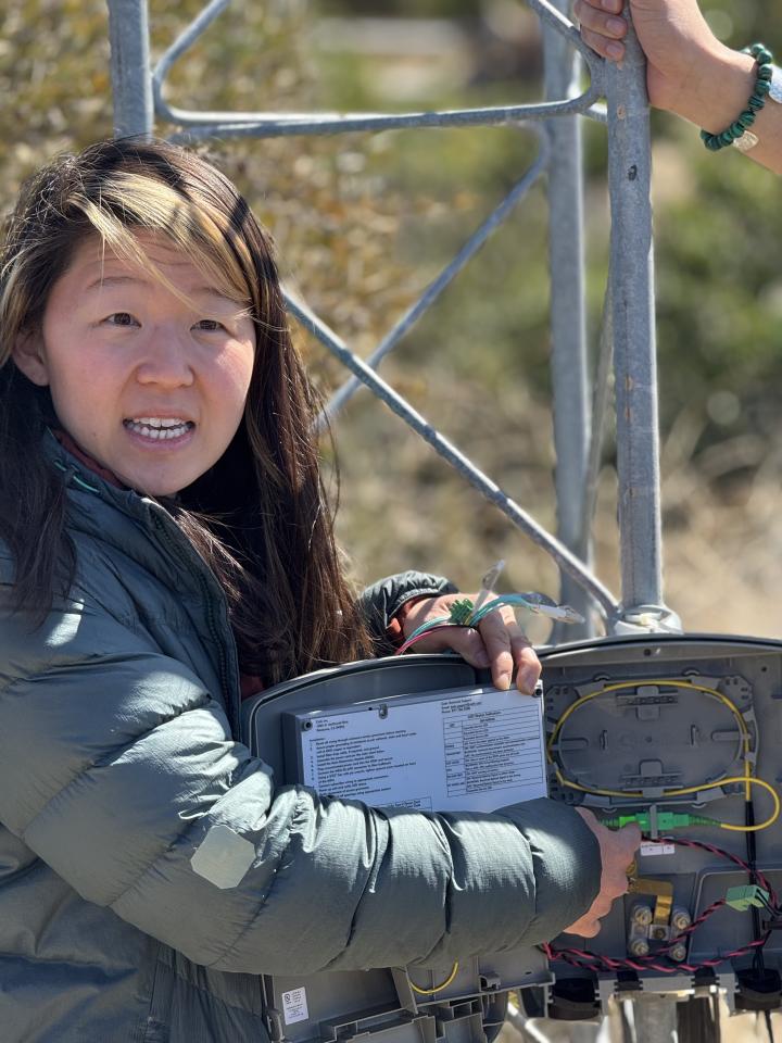 Esther at TBB 16 A woman in a green jacket holds open a wireless network access box at the base of a wireless tower